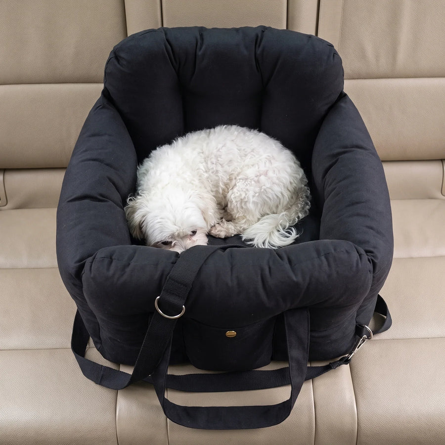 A small white Bichon curled up asleep inside the black Pawzie Perch booster bed on a car seat